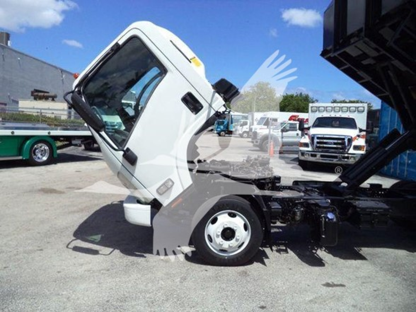 Isuzu truck cab tilted for maintenance in a lot; shows front wheel and chassis with dump bed in raised position.