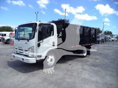 Isuzu NRR dump truck in view showing cab and dumping bed with some business lot in background.