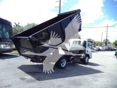 Isuzu dump truck in raised bed with black coating, side view showing hydraulic mechanism and cargo bed. No visible serials or labels legible.