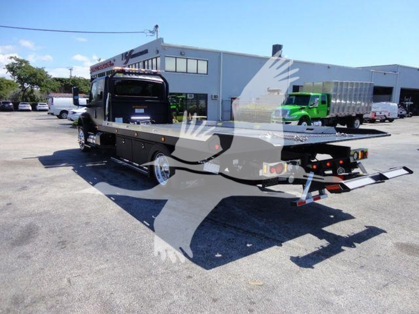 Truck bed rollback tow vehicle in a dealership lot with rear ramp, black cab, and green truck in background.