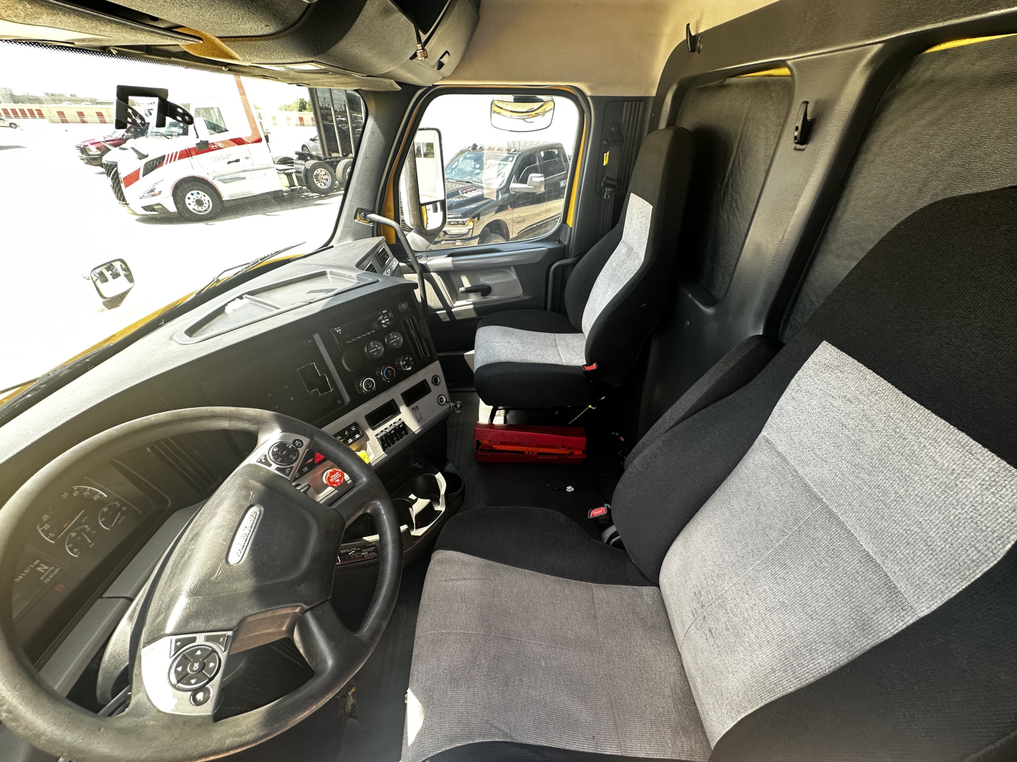 Dashboard and driver seat area of a 2019 Freightliner Cascadia 126 cab interior. Steering wheel, instrument cluster, center console, and passenger seat visible; gray fabric seats and black plastic trim.