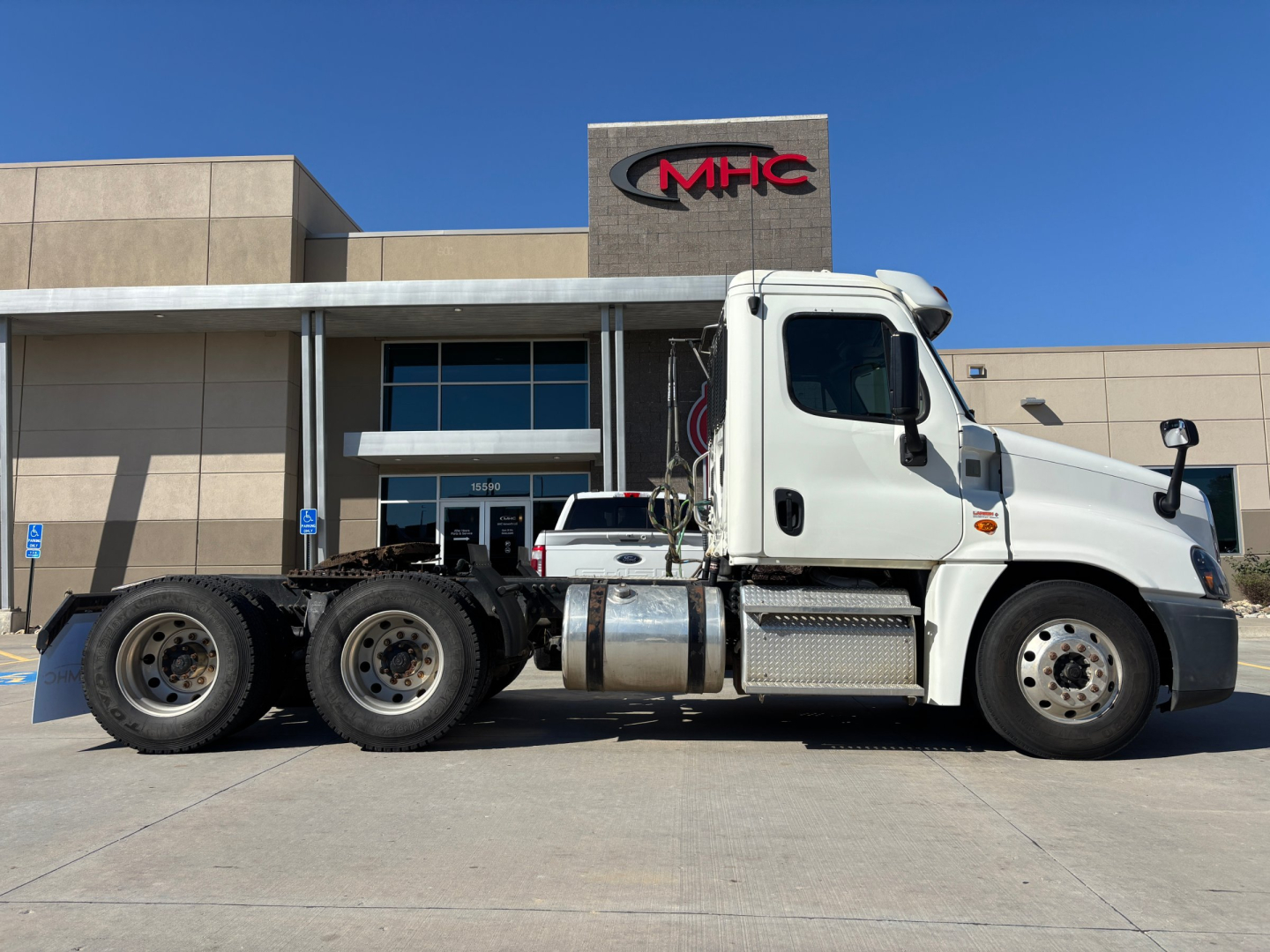 Truck side view of a white Freightliner CA12564DC conventional daycab tractor with dual rear axles and exposed fuel tanks parked in front of a building with MHC logo.