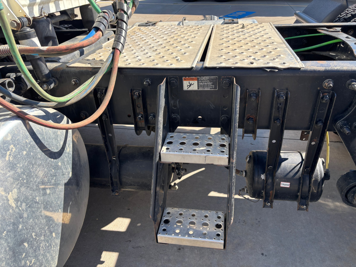 Conventional daycab trailer hitch area showing access steps and chassis with metal diamond-plate platform, hydraulic lines, and support brackets.