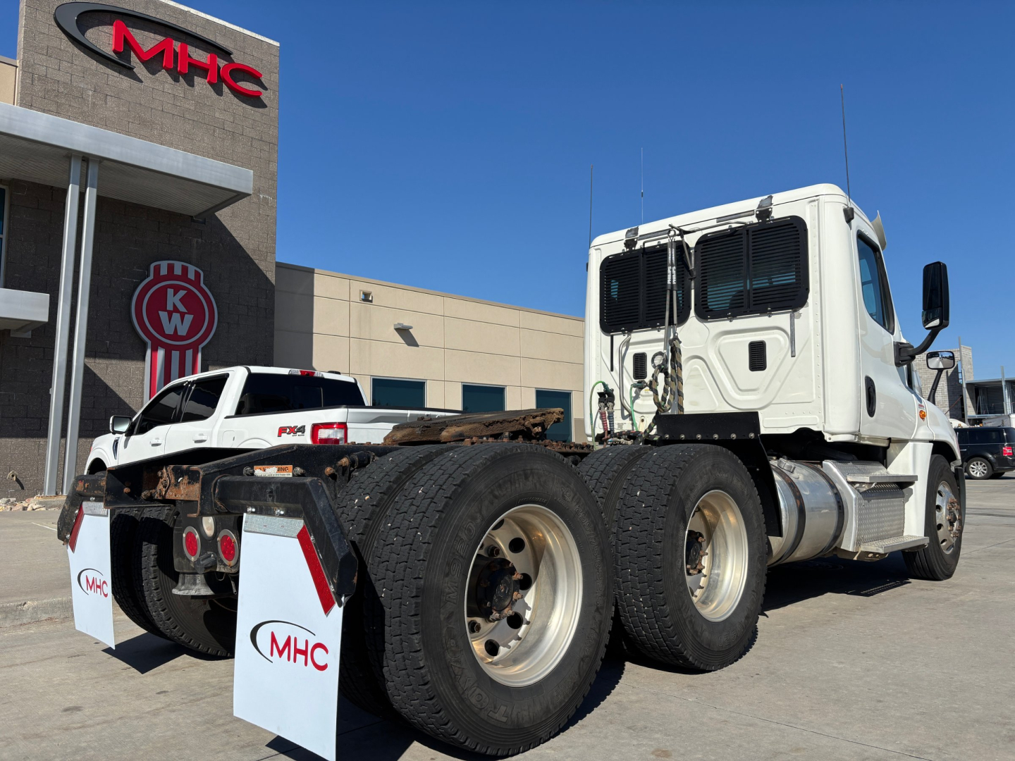Truck tractor in a dealership lot showing a Freightliner CA12564DC model with a white cab and dual rear axles, MHC branding on white wheel chocks, and a white service vehicle in background.