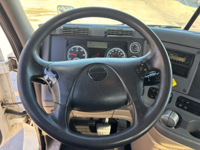Steering wheel of Freightliner CA12564DC cab with dashboard gauges visible behind; no visible text legible except circular gauge labels.