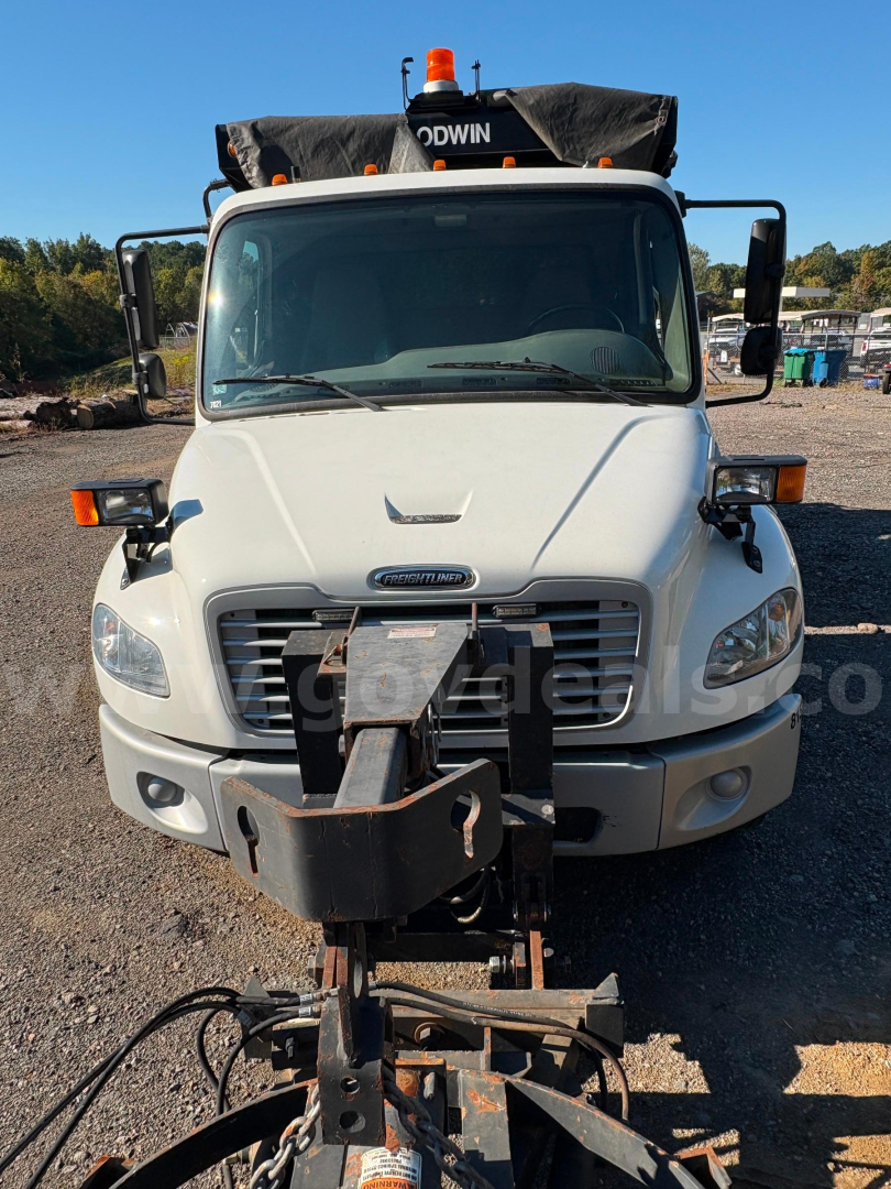 Front view of a 2015 Freightliner M2 dump truck showing a large front bumper with a mounted snowplow or attachment and amber beacon on cab roof.