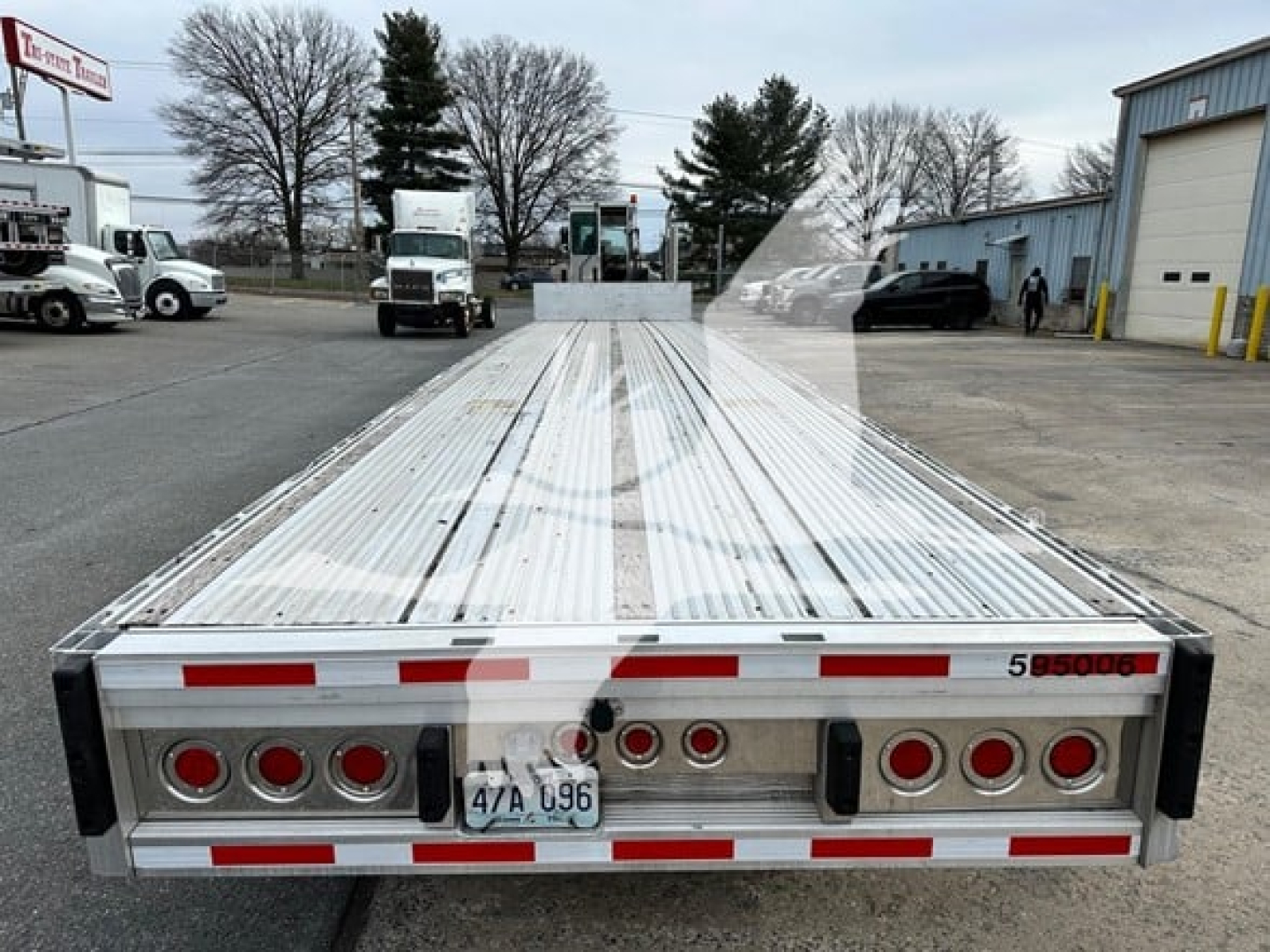 Flatbed trailer rear view showing multiple rectangular taillights, reflective tape, license plate, and rear bumper; aluminum bed with planks; compact tail assembly.