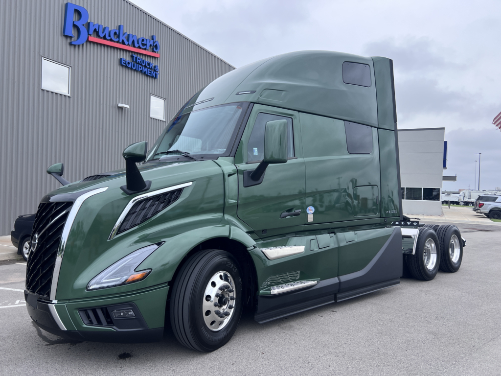 Green Volvo VNL64T860 cab with sleeper, front left view, showing curved hood, large side mirrors, and chrome wheel rims.