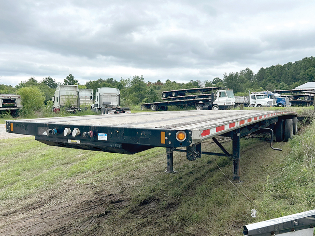Flatbed trailer showing electronic controls and metal deck with warning lights from the side, Fontaine flatbed with dual rear axles in a field.