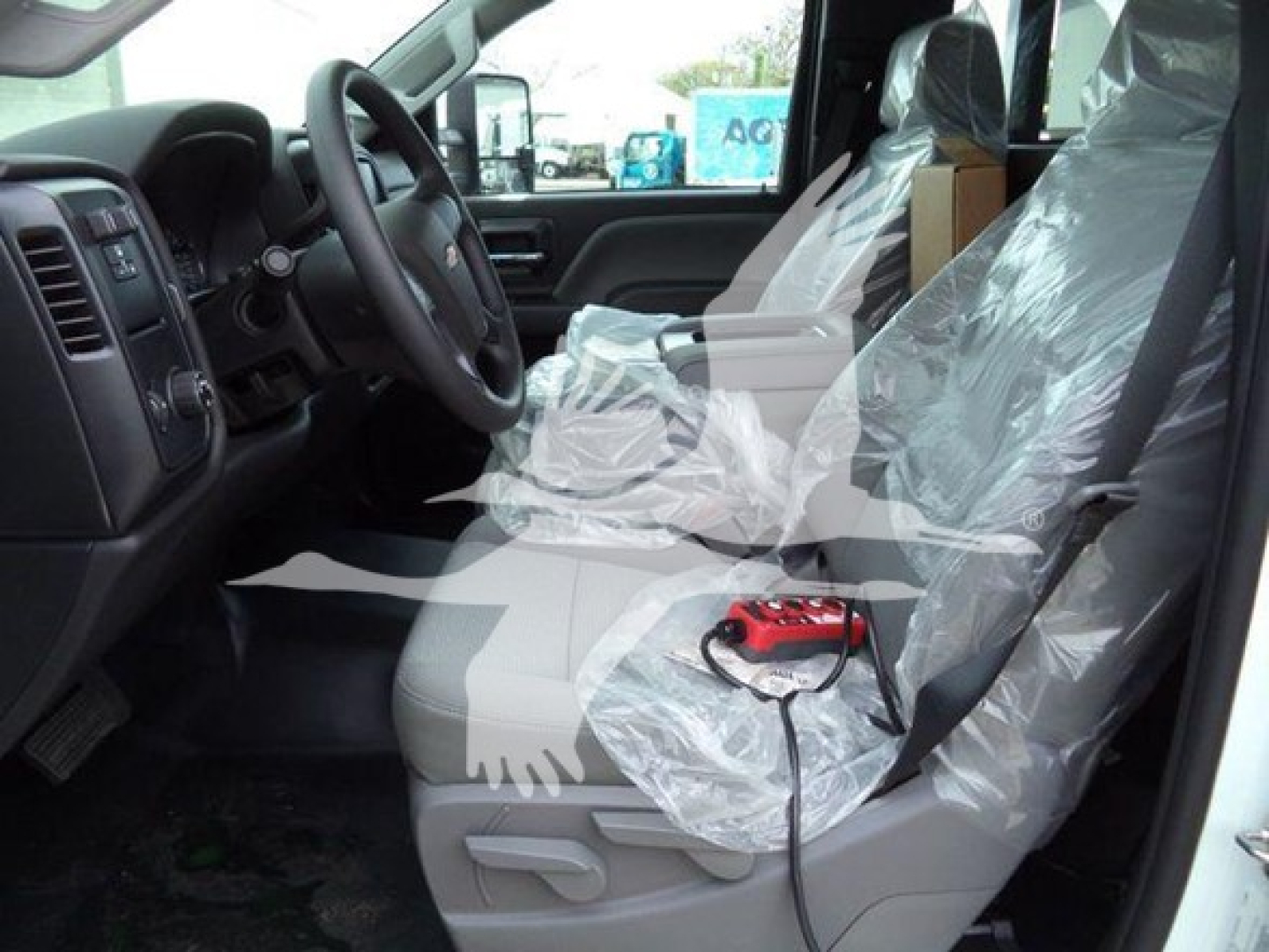 Interior of a new 2023 Chevrolet Silverado 6500HD cab with plastic seat covers and a red handheld device on the passenger seat; steering wheel and dashboard visible.