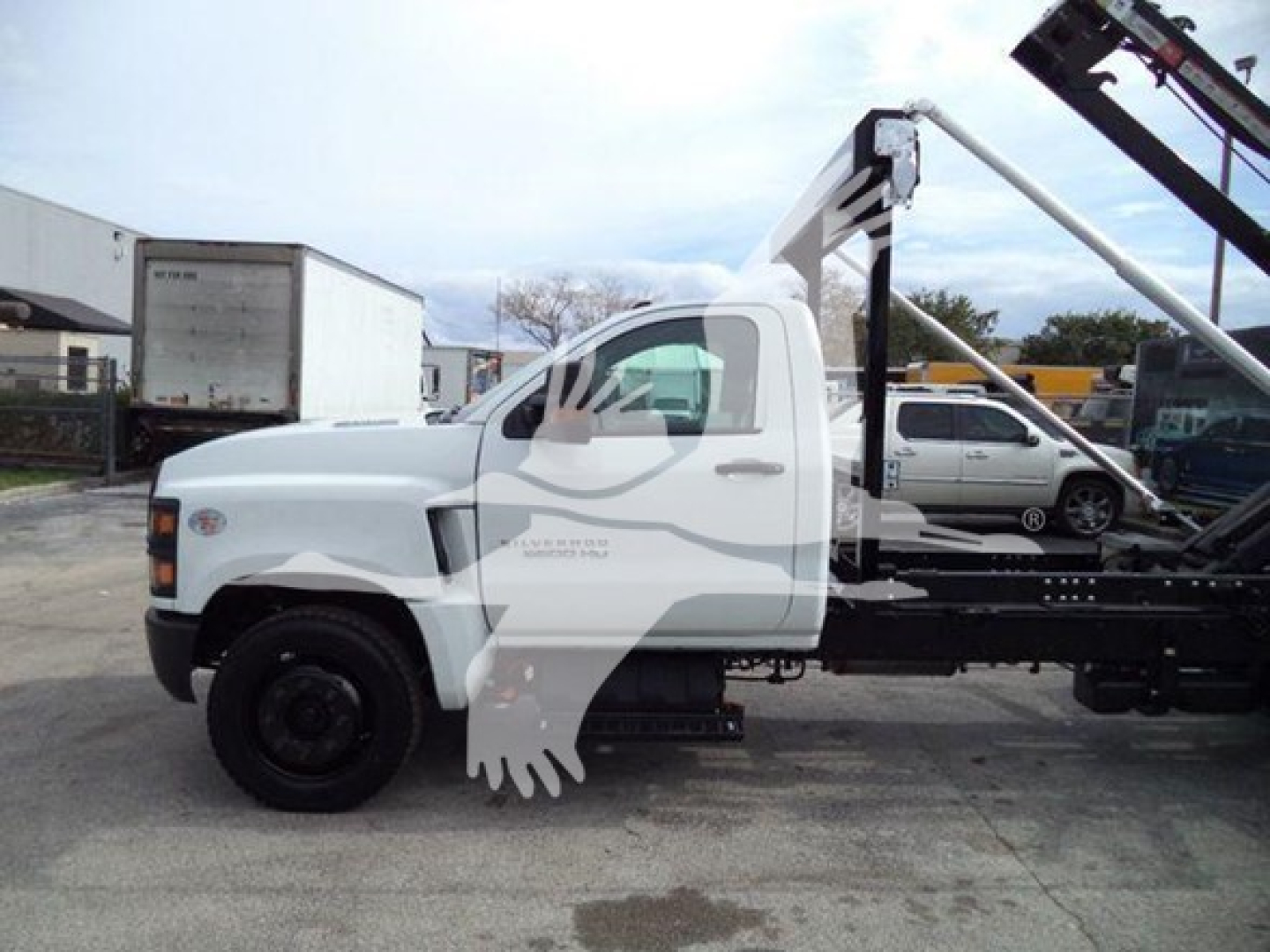 Truck side view showing a white 2023 Chevrolet Silverado 6500HD roll-off with flatbed and crane equipment.