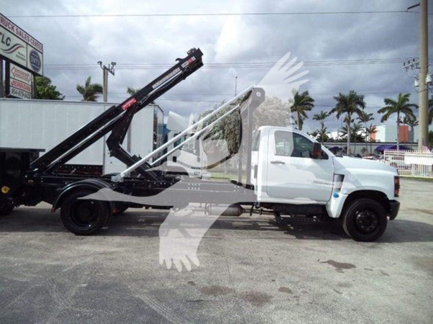 Truck with a long black loading arm on a white Chevrolet Silverado 6500HD chassis, side view, flatbed with roll-off hoist system.