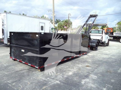Flatbed trailer with black cargo container and tow truck in background, roll-off category vehicle.