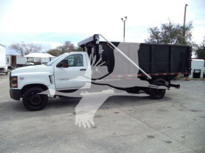 Truck in a lot showing a white Chevrolet Silverado 6500HD chassis cab with a black dump bed/roll-off container, side view, with a watermark overlay.
