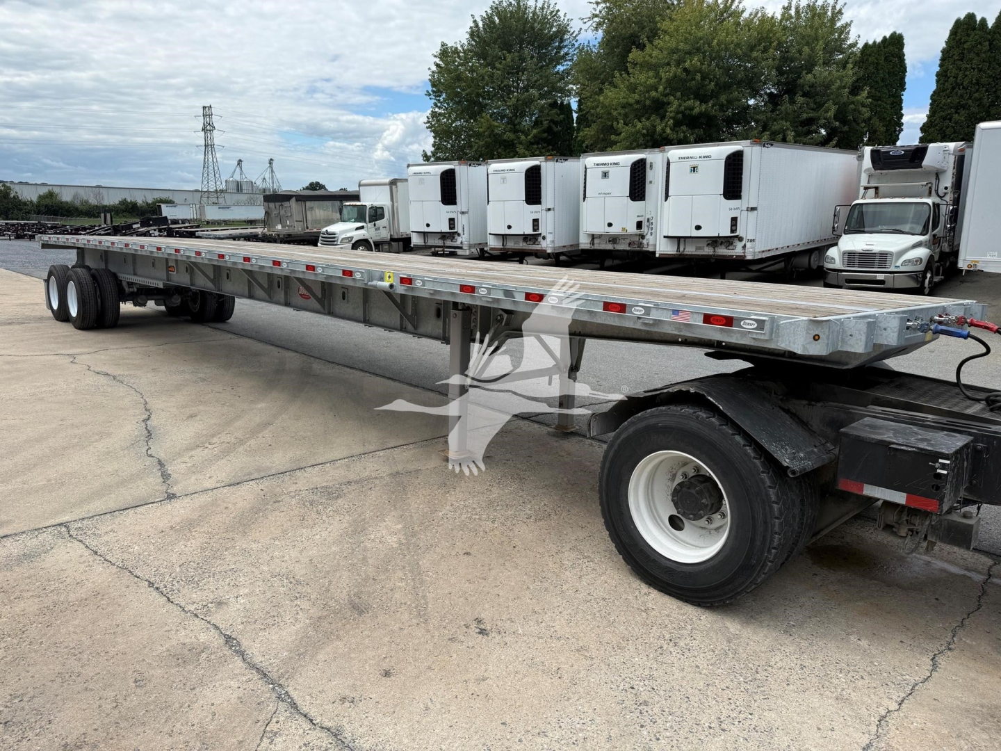 Flatbed trailer with extendable section showing long steel deck, dual rear axles, and cab of truck in distance; visible VIN tag and stock number VED49033; industrial yard with trailers in background.