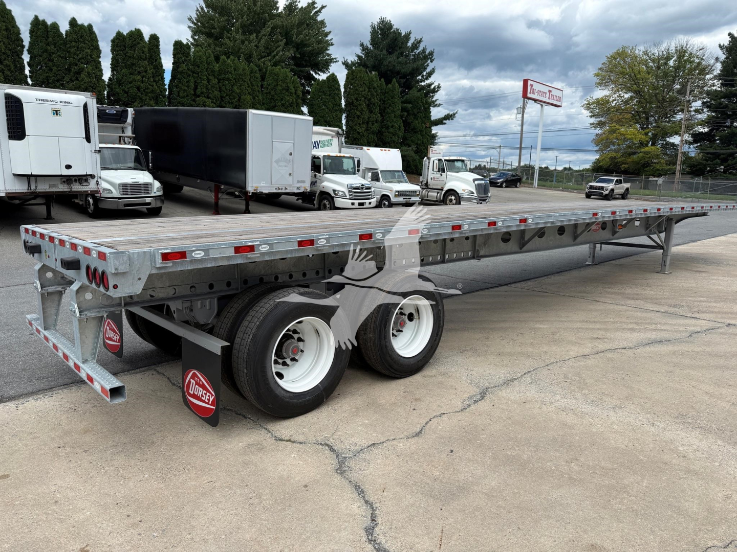 Flatbed trailer showing dual rear axles and galvanized steel bed with red/white reflective tape along sides; Dorsey logo mudflaps; industrial lot with trucks in background.