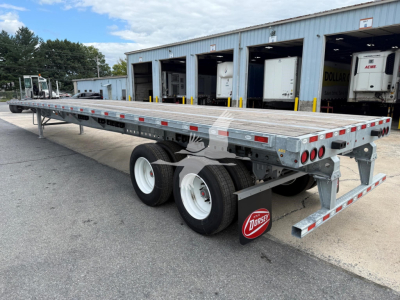 Flatbed trailer shown from rear-left side with dual rear axles, galvanized steel bed, rear underride guards, and Dorsey logo mudflap; DOT lights along sides; industrial warehouse background.