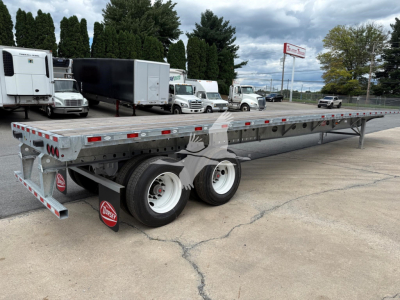 Flatbed trailer showing dual rear axles and galvanized steel bed with red/white reflective tape along sides; Dorsey logo mudflaps; industrial lot with trucks in background.