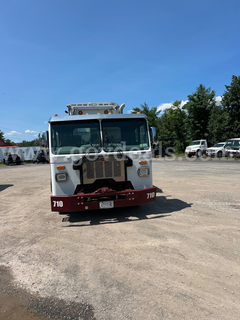 Peterbilt garbage truck front view showing white cab, red bumper with '710' numbers, and grille, in a lot with other trucks.