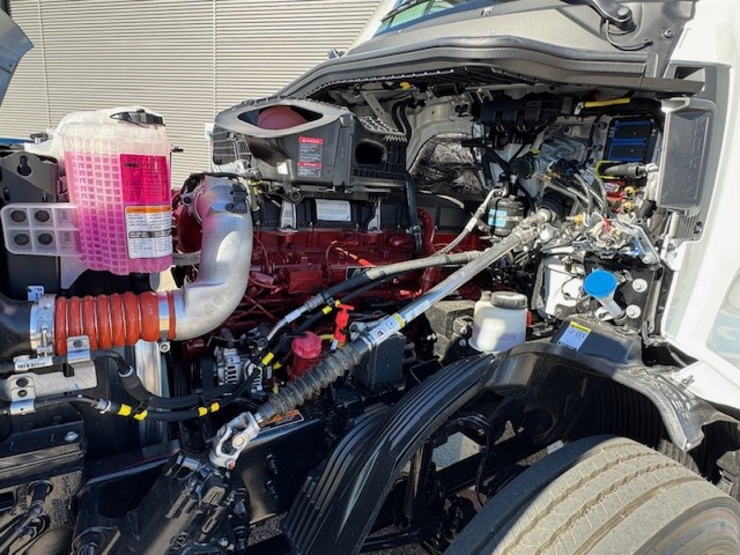 Engine bay of a 2026 Mack PR64T showing red engine block, pink coolant reservoir, various hoses, and wiring harnesses visible.