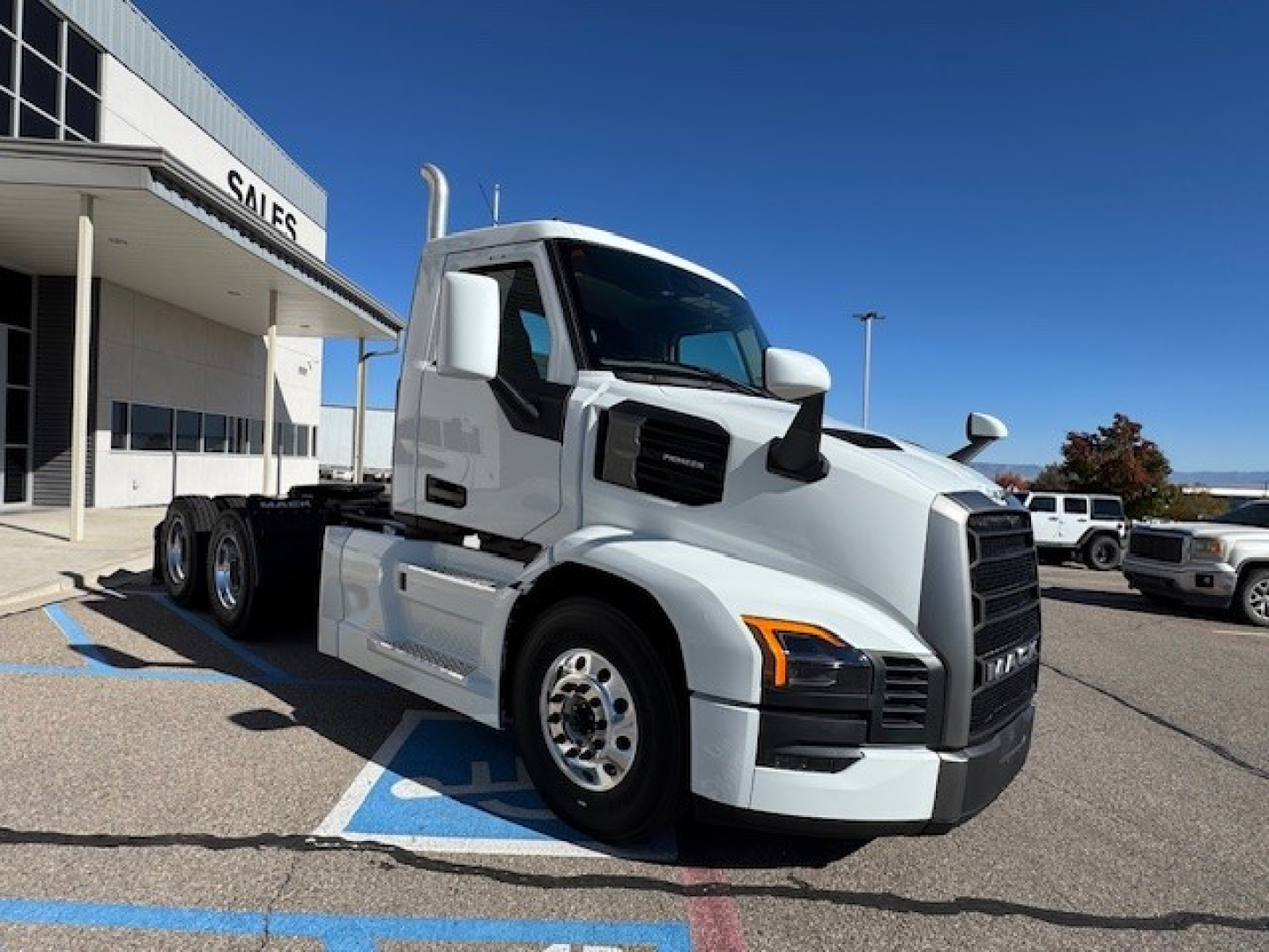 Truck cab front-left view showing Mack PR64T with white exterior, large black grille, and silver wheels; dealership building in background.