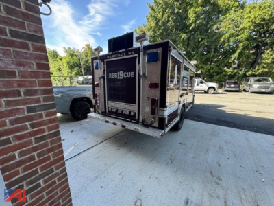Ambulance-style rescue vehicle or fire rescue truck with rear doors, text on back reading ‘RESQCRUE’ and license plate area, parking lot backdrop with trees and other vehicles.