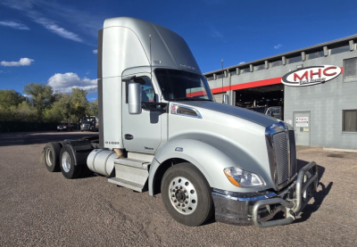 Kenworth T680 truck in front view showing cab and front bumper with chrome grille guard, silver paint, wheel rims, dual rear axles, and MHC dealership in background.