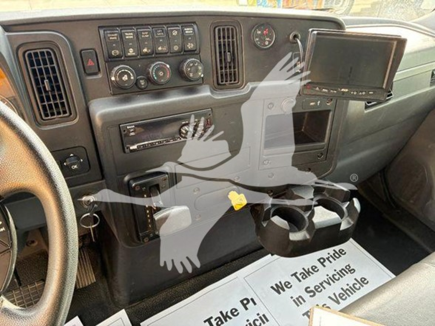 Interior view of a commercial truck cab dashboard showing controls, digital display, cup holders, and a hanging paper mat with text.