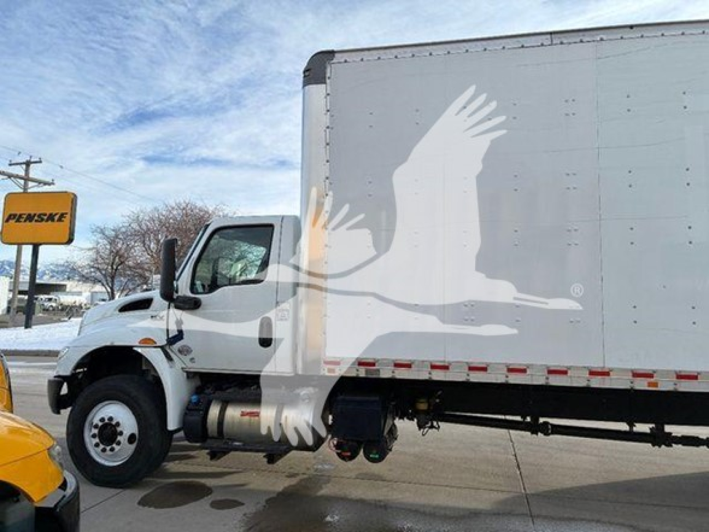 Truck cab and box van showing side view with white cargo box and black undercarriage. Visible text includes PENSKE sign in background; no readable technical specs on vehicle.