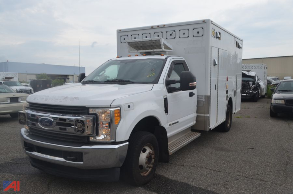 Ford F-350 Super Duty ambulance front-left view with box body, cab doors, and chrome bumper. Visible text: Ford logo on grille; amber clearance light bar on roof; small labels on ambulance box panels.