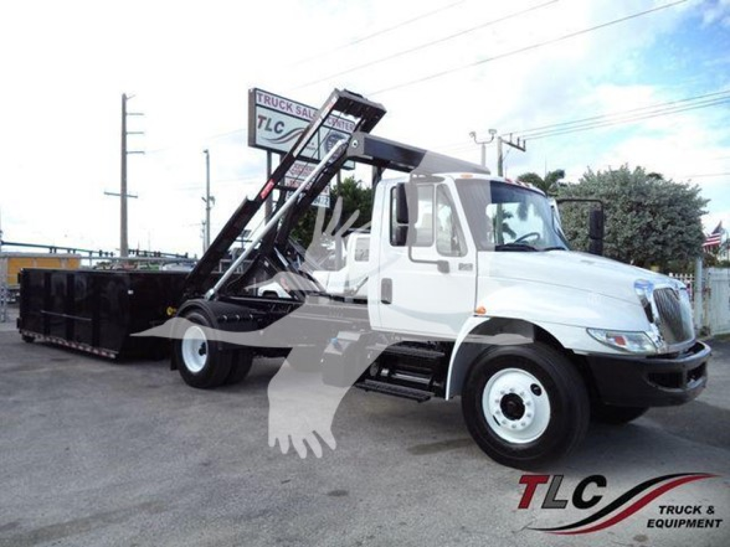 International Durastar 4300 roll-off truck shown from side angle with lifting arm and container, white cab, black bed, TLC signage faint in background