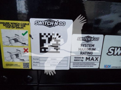 Sticker labels on a black surface showing SWITCH-N-GO system label, 15000 lb max, 6800 kg max, serial/model/part fields; left graphic with instructions for winch cable routing.