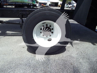Wheel and tire of a dump truck chassis with white rim and black tire; background shows trailer and yard equipment.