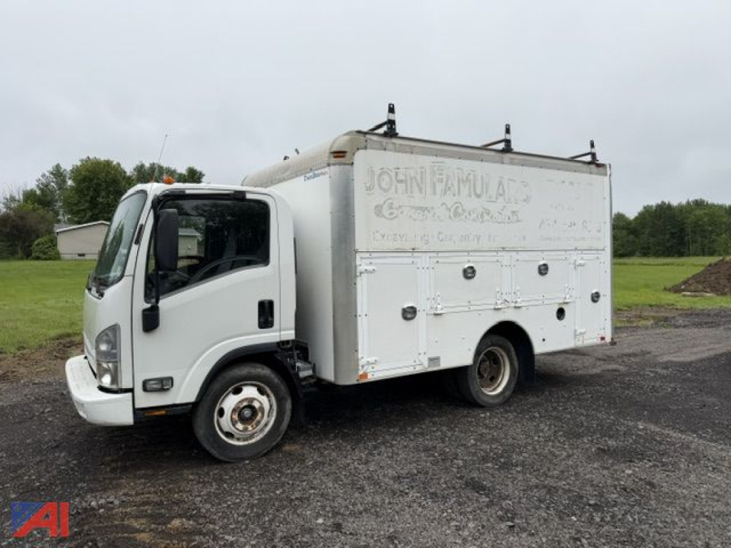 Isuzu NPR box truck with white service body showing faded lettering 'JOHN FAMULA' on side; cab front left; service compartments along side; exposed wheels and gravel lot.
