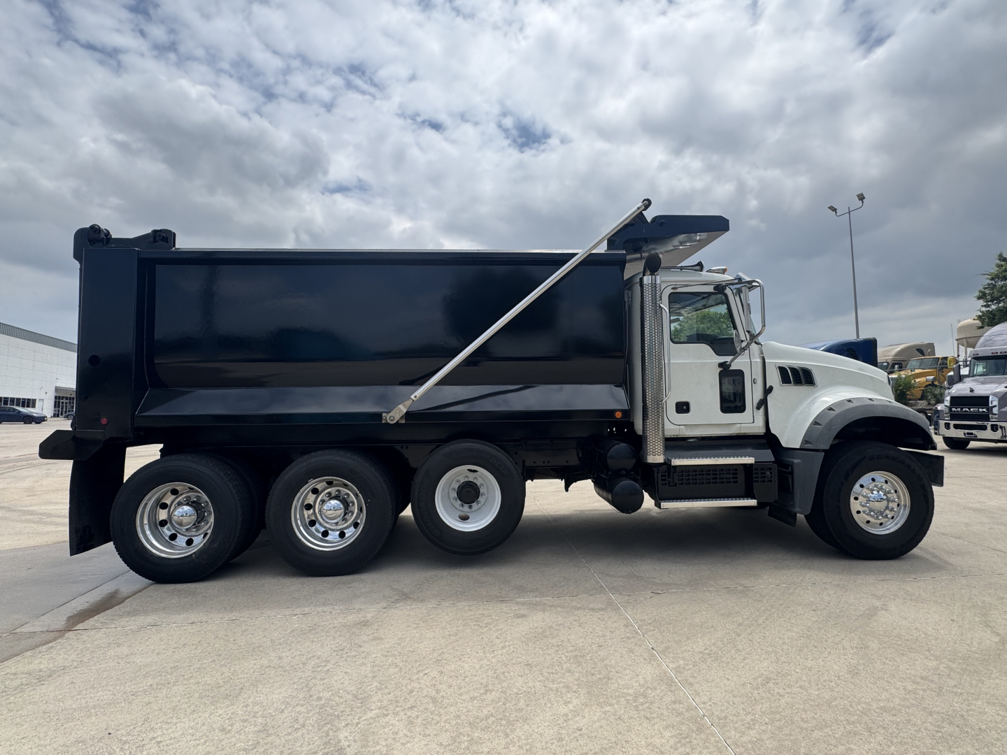 Dump truck side view showing 2021 Mack GR64F with black dump body, white cab, chrome wheels, and raised tarp bar.