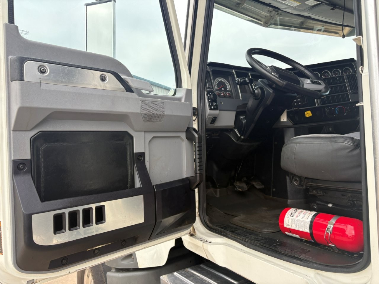 Cab interior of 2021 Mack GR64F dump truck showing open door, dashboard, steering wheel, and fire extinguisher on floor near pedals.