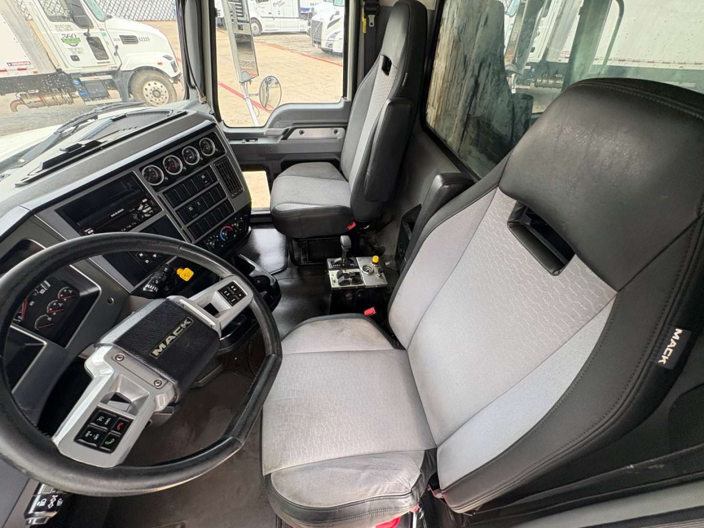Mack GR64F truck cab interior showing steering wheel, dashboard controls, and driver seat with light gray fabric.