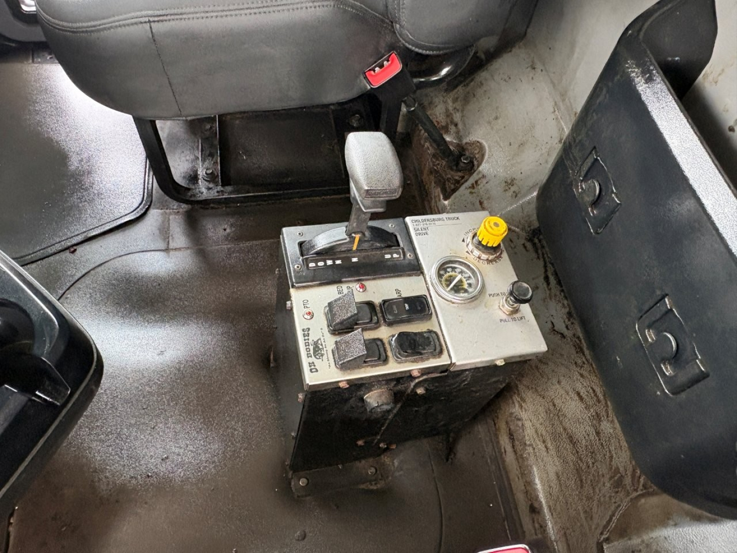 Cab interior showing pedal area and a rectangular control console with levers, buttons, and a yellow cap; seat and floor area visible; fluid reservoir and switches on panel.