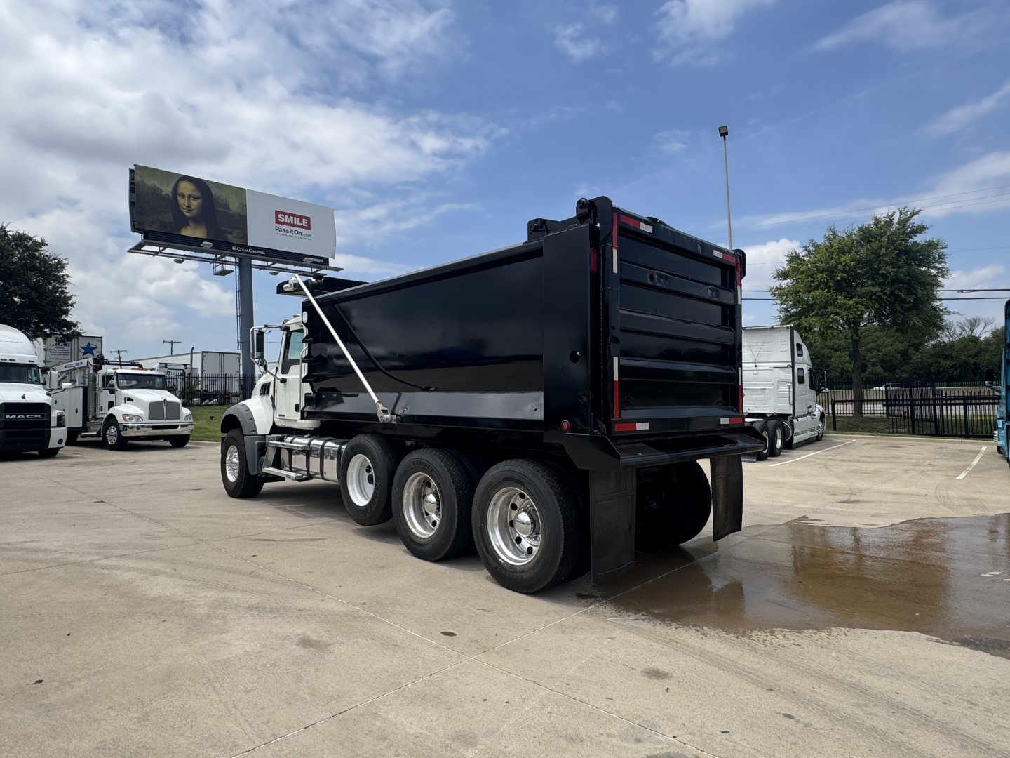 Dump truck with black dump body and tandem rear wheels. Visible text on side of cab and near rear tailgate; parking lot with a billboard in background.