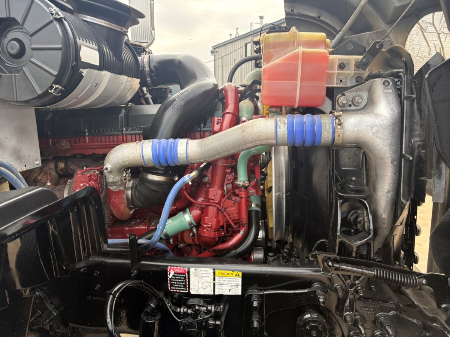 Mack GR64F engine bay showing red engine block, blue hose clamps, coolant reservoir, and turbo piping.