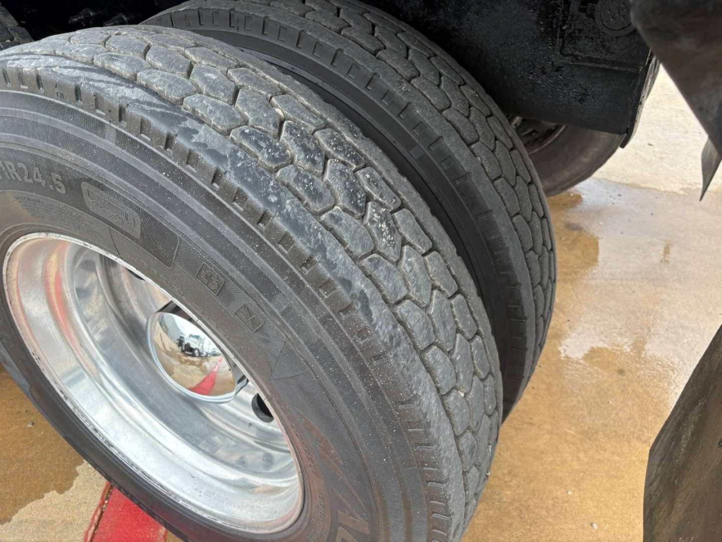 Truck wheel and dual rear tires visible; tread pattern and polished steel rim; slight mud and water on ground.