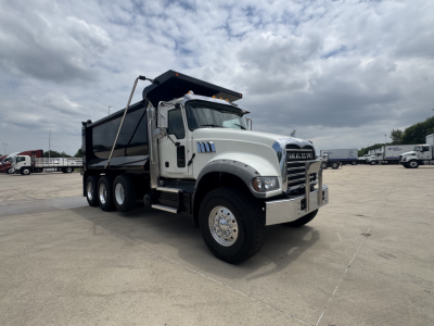Truck image showing a white 2021 Mack GR64F dump truck with six wheels and a raised dump bed in a parking lot.