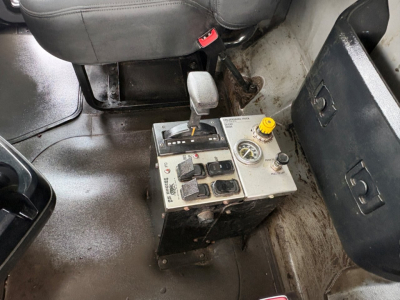Cab interior showing pedal area and a rectangular control console with levers, buttons, and a yellow cap; seat and floor area visible; fluid reservoir and switches on panel.