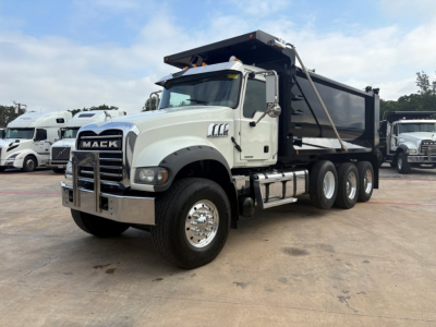 Mack GR64F dump truck in-service showing white cab and black dump bed with raised tarp bars.
