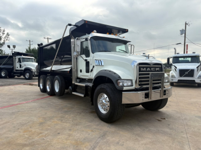 Mack dump truck in a lot, front three-quarter view showing the Mack grille, bumper, and dump bed. Visible text includes 'MACK' on the grille badge and no other legible labels.