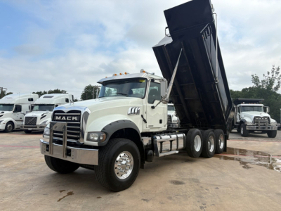 Mack GR64F dump truck with raised dump bed in a dealership lot, white cab, chrome bumper, dual rear axles, and MACK emblem on the grille.