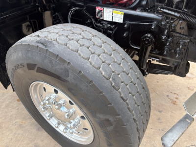 Front left wheel and undercarriage of a Mack GR64F dump truck showing heavy-duty tire, chrome wheel, and suspension/components. Visible labels on frame include small warning stickers and labels.