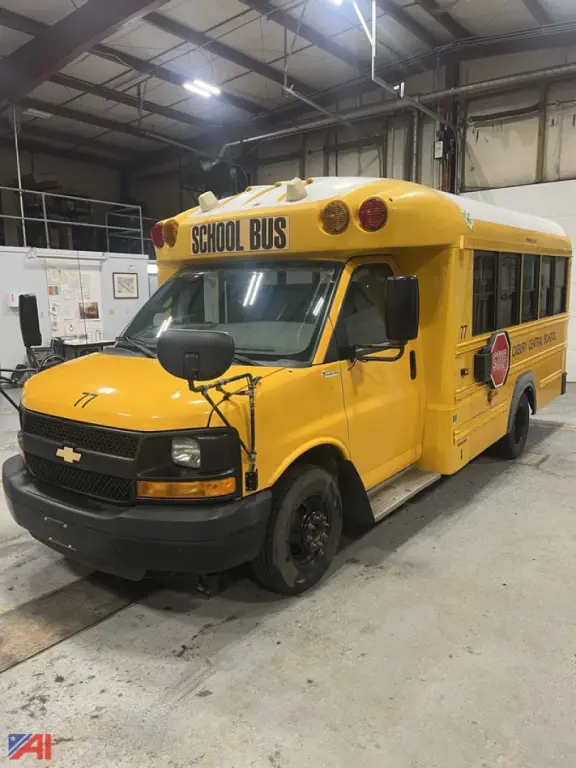 School bus in a garage, 2016 Chevrolet Express, yellow body, front view with STOP sign, black bumper and wheels, 'SCHOOL BUS' signage on top front and side