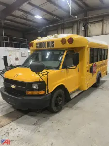 School bus in a garage, 2016 Chevrolet Express, yellow body, front view with STOP sign, black bumper and wheels, 'SCHOOL BUS' signage on top front and side