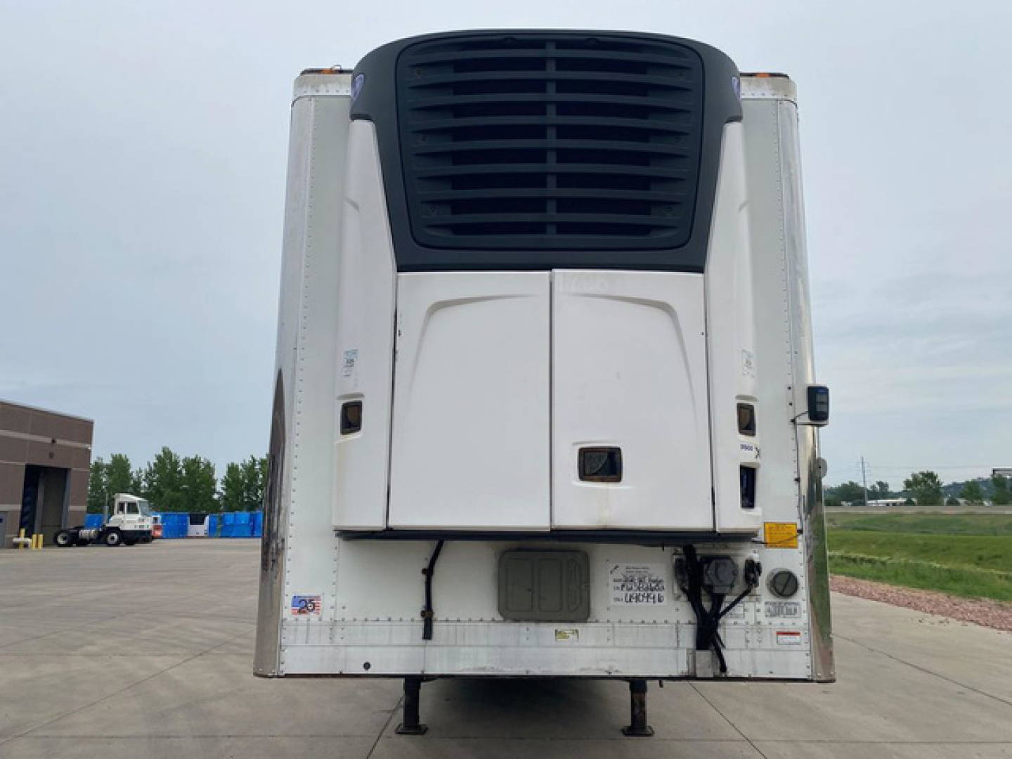 Refrigerated trailer rear showing cargo unit with black upper ventilation grill, white doors, and undercarriage legs; VIN sticker area and miscellaneous labels visible.